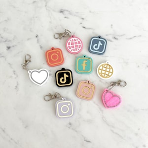 Może przedstawiać: Kolekcja kolorowych breloków z motywem mediów społecznościowych. Breloki przedstawiają różne ikony, w tym Instagram, TikTok, Facebook i globus. Każdy brelok ma srebrne zapięcie i jest w odcieniach różu, pomarańczy, błękitu i złota.
