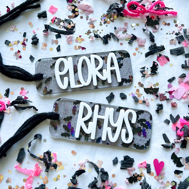Pode incluir: Duas etiquetas de nome personalizadas com fundos de glitter preto e branco. Os nomes "Elora" e "Rhys" est&atilde;o escritos em branco nas etiquetas. As etiquetas est&atilde;o presas a cord&otilde;es pretos.