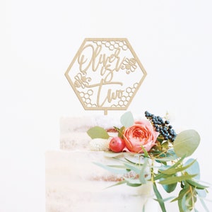 May include: Wooden cake topper in the shape of a hexagon with a honeycomb pattern. The topper says "Oliver is two" in a handwritten font with two bees on the side.