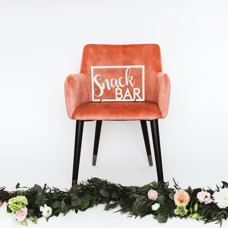 Puede incluir: Una silla de terciopelo rosa coral con patas negras se encuentra frente a una pared blanca. Un letrero de madera blanco con el texto "Snack Bar" se coloca encima de la silla. Una guirnalda de vegetaci&oacute;n y flores se encuentra frente a la silla.