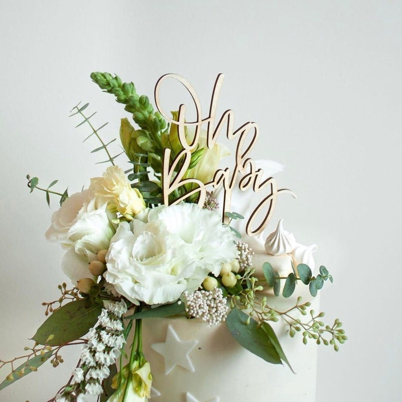 May include: White cake with a wooden "Oh Baby" topper and white flowers. The cake is decorated with white stars.
