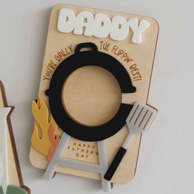 May include: Wooden Father's Day card with a grill, flames, and a spatula. The card says "Daddy, You're Grilly The Flippin' Best!" and "Happy Father's Day".
