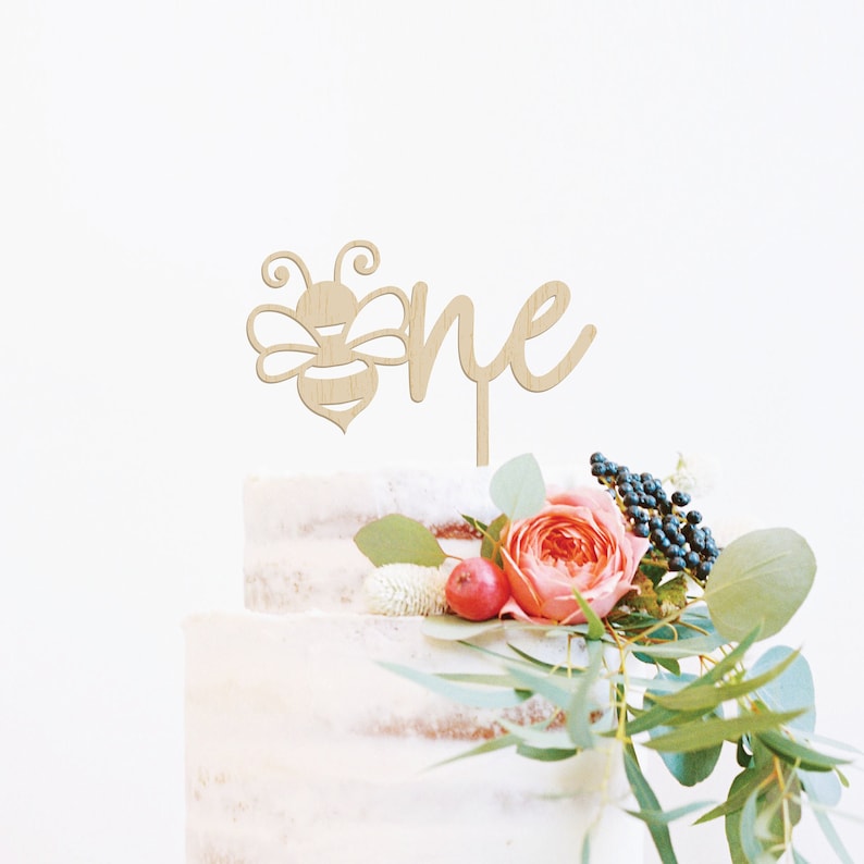 May include: A wooden cake topper featuring a bee design and the word "one". The topper is light brown and is placed on a two-tiered cake decorated with flowers and greenery. The cake has a rustic, semi-naked appearance.