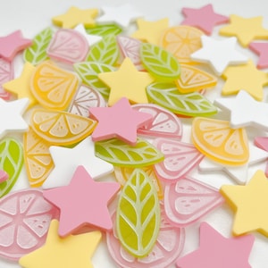 May include: A selection of colourful craft embellishments, featuring star shapes in white, pink, and yellow, alongside translucent green leaves and citrus fruit slices in pink and orange. These decorative pieces are scattered across a white surface.