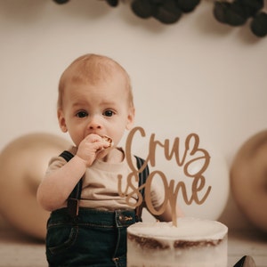 Puede incluir: Un beb&eacute; con un peto vaquero y una camiseta beige, comiendo un bocadillo. Delante del beb&eacute; hay una tarta con una decoraci&oacute;n de madera que dice "Cruz is One". Globos dorados y blancos en el fondo.