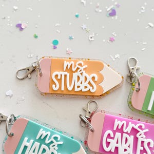 Pode incluir: Quatro chaveiros em forma de l&aacute;pis coloridos com detalhes de glitter. Cada chaveiro tem o nome de um professor diferente: "Ms. Stubbs", "Ms. Harris", "Mrs. Garbles" e "Mrs. [nome ileg&iacute;vel]".