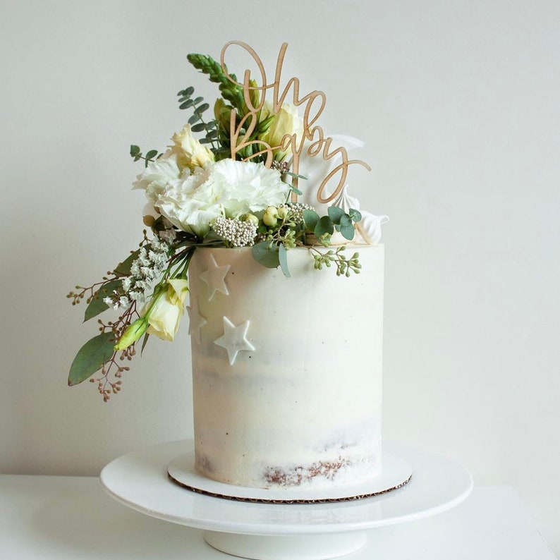 May include: A white cake with a light brown bottom layer decorated with white stars and topped with white flowers and greenery. A wooden cake topper with the words "Oh Baby" is placed on top of the cake.