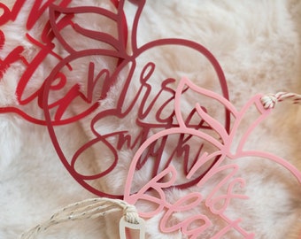May include: Three red and pink apple-shaped ornaments with the text "Mrs. Smith" and "Mrs. Beatty" on them. The ornaments are hanging from a white fuzzy surface.