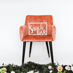 Puede incluir: Una silla de terciopelo de color coral con patas negras y un letrero de madera blanco que dice "Snack Bar" en la parte superior.