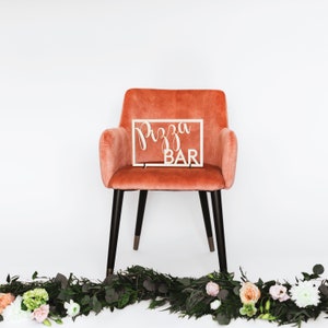 Puede incluir: Una silla de terciopelo rosa con patas negras y un letrero de madera que dice "Pizza Bar" en la parte superior.