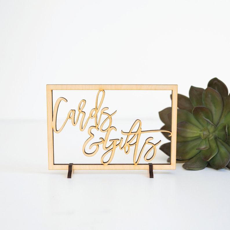 Pu&ograve; includere: Un cartello in legno con le parole "Cards & Gifts" in un font corsivo. Il cartello &egrave; incorniciato da un sottile bordo in legno e ha due piccole gambe in legno.
