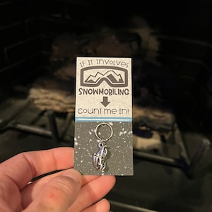 May include: A snowmobile keyring with a silver-toned charm, attached to a card. The card reads "IF IT involves SNOWMOBILING count me IN!" in black text, with a speckled background and a blue and white striped border.