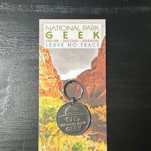 May include: A silver keychain with a round metal charm that says "National Park Geek" with a ranger hat and glasses graphic. The keychain is displayed on a card with the text "National Park Geek Explore. Discover. Adventure. Leave No Trace" and a watercolor image of a canyon.
