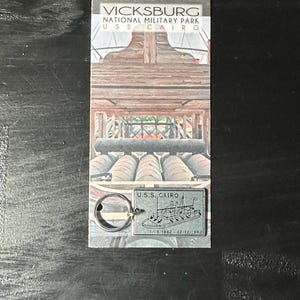 May include: A keychain featuring a black and white illustration of the U.S.S. Cairo, a Civil War ironclad. The keychain is attached to a silver key ring. The text on the keychain reads "U.S.S. CAIRO 1/16/1862 - 12/12/1862".