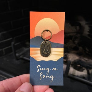 May include: A bronze key chain with a small oval charm featuring a stylized deer with a crescent moon and stars. The charm is attached to a silver key ring. The key chain is displayed on a card with a sunset background and the text "Sing a Song".