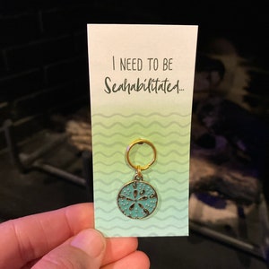 May include: A teal blue sand dollar keychain with a gold ring and a white card that says "I need to be Seahabilitated..." with a green and white wavy background.