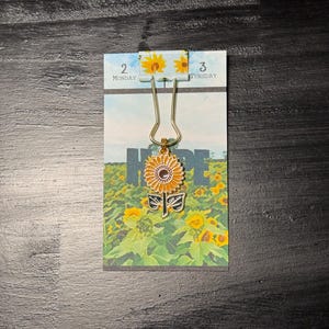May include: A gold-colored paper clip holding a small calendar page with the word "HOPE" and a sunflower charm. The charm features a yellow sunflower with a black and white butterfly design below it. The calendar shows the days Monday and Tuesday.