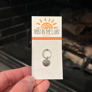 May include: A silver charm with a sun design and the words "TRUST IN THE LORD" and "Proverbs 3:5-6" on a white card.
