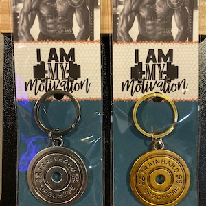 May include: Two silver and gold keychains with a weight plate design. The keychains feature the text "I am my motivation" and "Train Hard" with a 50 kg weight plate design.