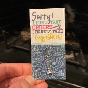May include: A silver keychain with a gavel charm. The keychain is packaged on a blue and white patterned card with the text "Sorry! I don't take orders I barely take suggestions".