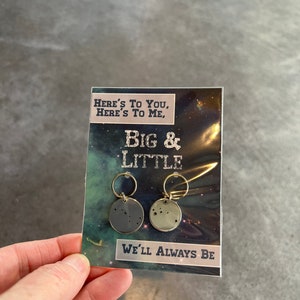 May include: A card with the text "Here's to you, Here's to me, Big & Little, We'll always be" and two small round silver charms with constellations on them.