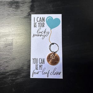 May include: A penny keychain with a four-leaf clover charm attached to a penny. The card reads "I can be your lucky penny, you can be my four-leaf clover" with a blue heart balloon graphic.