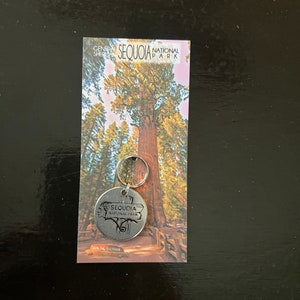 May include: A silver keychain with a round metal charm featuring the text "Sequoia National Park" and a stylized image of a tree. The keychain is attached to a card with a photo of a giant sequoia tree and the text "General Sherman Sequoia National Park".