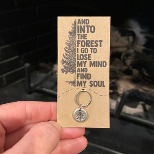 May include: A brown card with the text "And into the forest I go to lose my mind and find my soul" printed on it. A silver charm with a tree design is attached to a silver key ring.