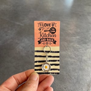 May include: A silver keychain with a frying pan charm featuring a white egg with a yellow yolk. The keychain is attached to a card with the text "I love you to the kitchen and back".