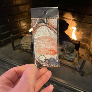 May include: A silver keychain with a quote by Vincent van Gogh: "Great things are done by a series of small things brought together." The keychain is in a clear plastic bag with a wood grain background.