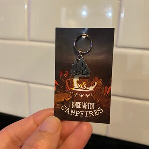 May include: A silver keychain featuring a black and silver campfire design. The keychain has a ring attached. The card behind the keychain reads "I Binge Watch Campfires".