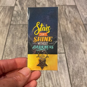 May include: A gold star-shaped charm with a ring attached. The charm has a dark blue background with a starry sky design. The text on the charm reads "Stars Can't Shine Without Darkness".