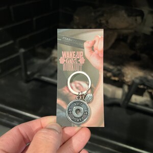 May include: Silver keychain with a weight plate charm that says "Lift Heavy" and a smaller charm that says "Strength". The keychain is attached to a card with the text "Wake Up and Workout".