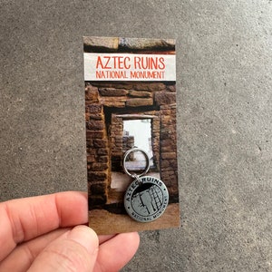 May include: A silver keychain with a circular design featuring the text "Aztec Ruins National Monument" and a depiction of a stone wall. The keychain is attached to a card with the text "Aztec Ruins National Monument" and a photo of a stone archway.