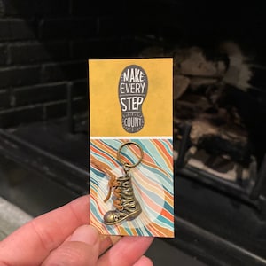 Peut inclure: Un porte-clés en forme de botte en cuir marron avec un charme en métal doré. Le charme porte les mots "Make Every Step Count".
