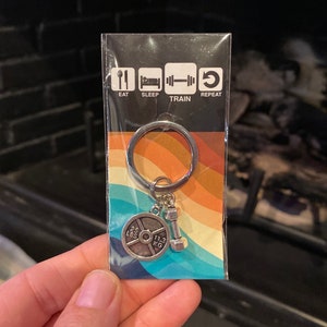 May include: A silver keyring with a weight plate charm that says "25 LBS 11.3 KG" and a dumbbell charm. The keyring is packaged on a card with the words "Eat, Sleep, Train, Repeat" and icons for each word.