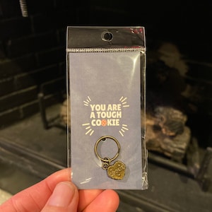 May include: A bronze-colored keychain with a heart-shaped charm that says "Tough Cookie". The keychain is packaged in a clear plastic bag with a blue card that says "You are a tough cookie".