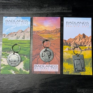 Könnte beinhalten: Drei Schlüsselanhänger vom Badlands National Park. Jeder Schlüsselanhänger hat ein anderes Design: einen Bison, einen Dickhornschaf und einen Präriehund. Die Schlüsselanhänger sind an Karten mit Landschaftsillustrationen und dem Parknamen befestigt.