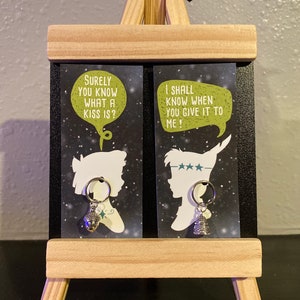 May include: Two black cards with white silhouettes of Peter Pan and Wendy. The cards have speech bubbles with text from the movie Peter Pan. Each card has a silver acorn charm attached to a silver ring.