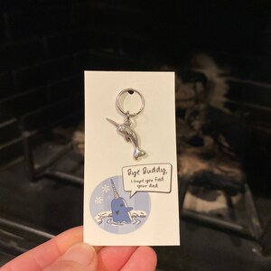May include: A silver narwhal keychain with a blue and white cartoon narwhal on a white card with the text "Bye Buddy, I hope you find your dad".