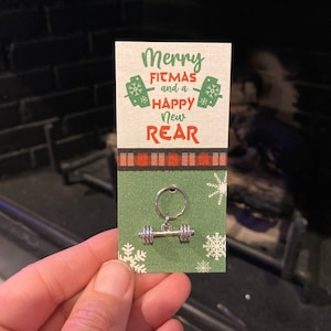 May include: A green and red card with the text "Merry Fitmas and a Happy New Rear" and a silver dumbbell keychain attached.