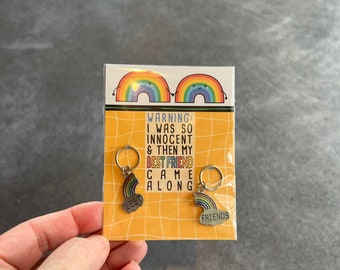 May include: Two rainbow-colored keychains with a cloud-shaped charm. The keychain on the left says "Best" and the keychain on the right says "Friends". The card behind the keychains says "Warning: I was so innocent & then my best friend came along".