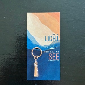 Könnte beinhalten: Goldfarbener Schlüsselanhänger mit einem Leuchtturm-Anhänger mit den Worten "be a light for all to see" und "matthew 5:16" auf blauem und orangefarbenem Hintergrund.