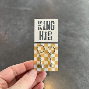 May include: A white card with black text that reads "HER QUEEN HIS KING" with a chess piece graphic. Two silver keychains with chess piece charms are hanging from the card.