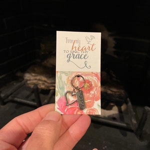 May include: A silver charm with a heart and the word "GRACE" on a tag. The charm is attached to a key ring. The charm is on a card with a floral design and the text "Tune my heart to sing thy grace".
