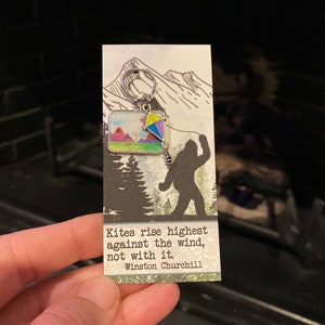 May include: A silver keychain with a colorful kite and mountain scene charm. The keychain has a quote by Winston Churchill: "Kites rise highest against the wind, not with it."