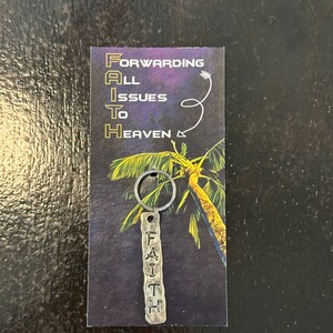 Könnte beinhalten: Ein silberner Schlüsselanhänger mit einem Palmenmotiv und dem Wort "FAITH" auf einem Metallanhänger. Der Schlüsselanhänger hat einen kleinen Ring. Der Hintergrund ist eine Postkarte mit dem Text "FORWARDING ALL ISSUES TO HEAVEN".