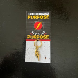May include: A gold lightning bolt keychain with a ring. The keychain is packaged on a white card with the text "Life doesn't give us purpose. We give life purpose."