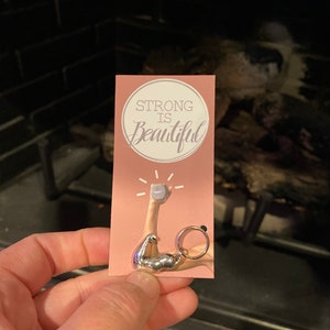 May include: A pink card with the text "STRONG IS Beautiful" in a circle. A silver charm of a bicep lifting a small dumbbell is attached to a silver ring.