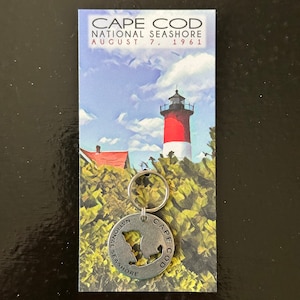 May include: A silver keychain with a cutout of Cape Cod and the text "National Seashore" around the edge. The keychain is on a postcard with a lighthouse and the text "Cape Cod National Seashore August 7, 1961".